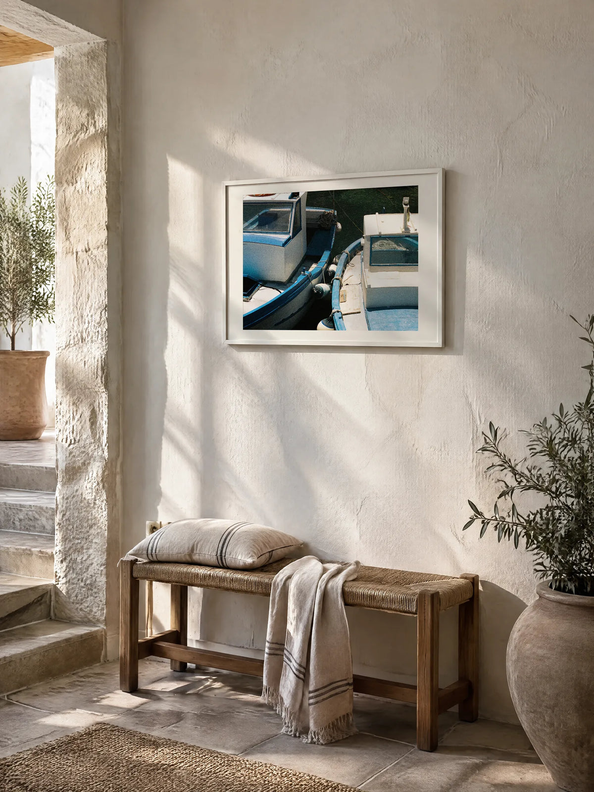 Blue Boats by Laís Tomaselli, framed in white oak with a white mat (24 × 18 in), above a bench in a Mediterranean entryway